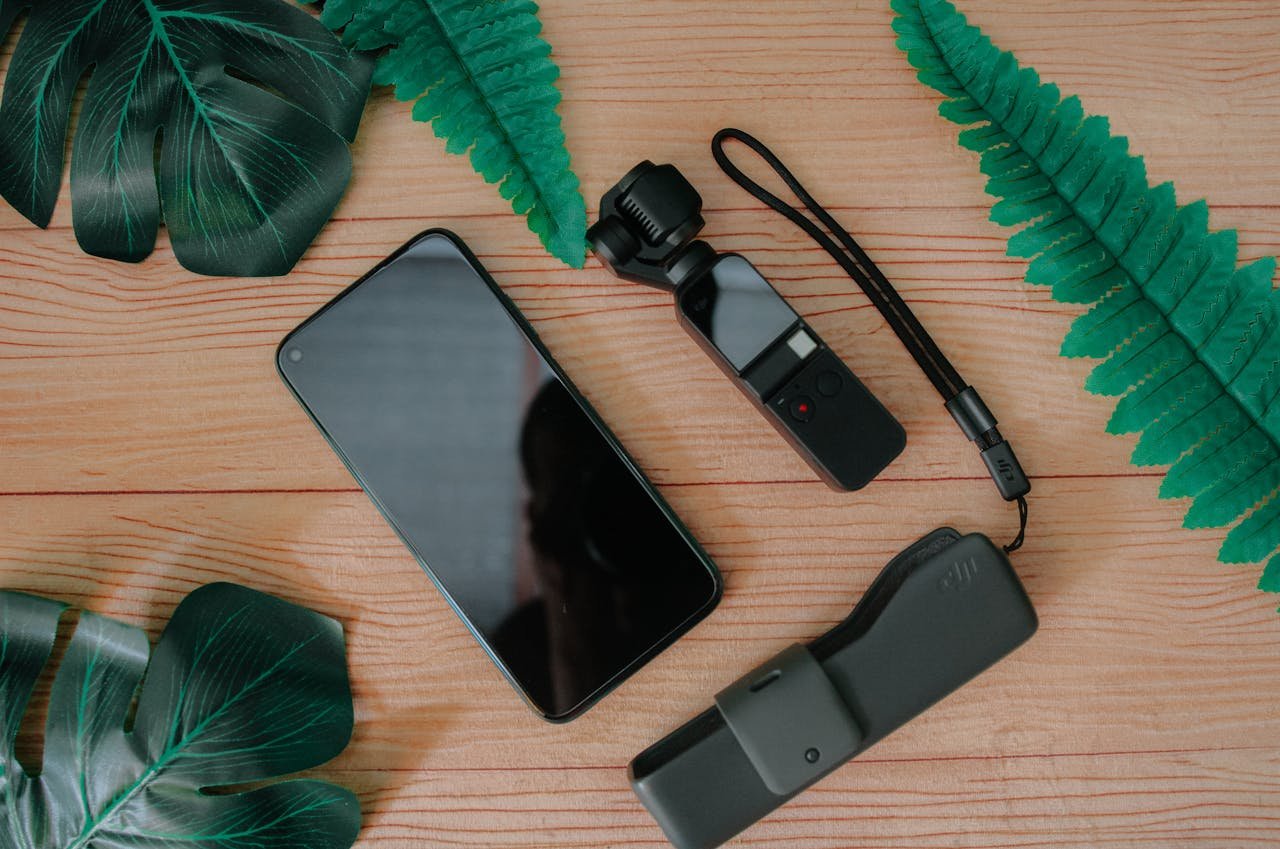 gallery-img-04 Top view of mobile phone and mini handheld camera with flash drive composed on wooden table with green leaves of tropical plants