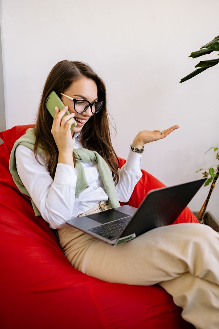 Smiling woman with glasses on a bean bag chair using a laptop while talking on the phone.